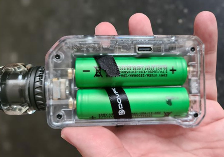 A hand holding the transparent version of the Dovpo MVP mod, showing the two 18650 batteries clearly visible through the clear outer shell.