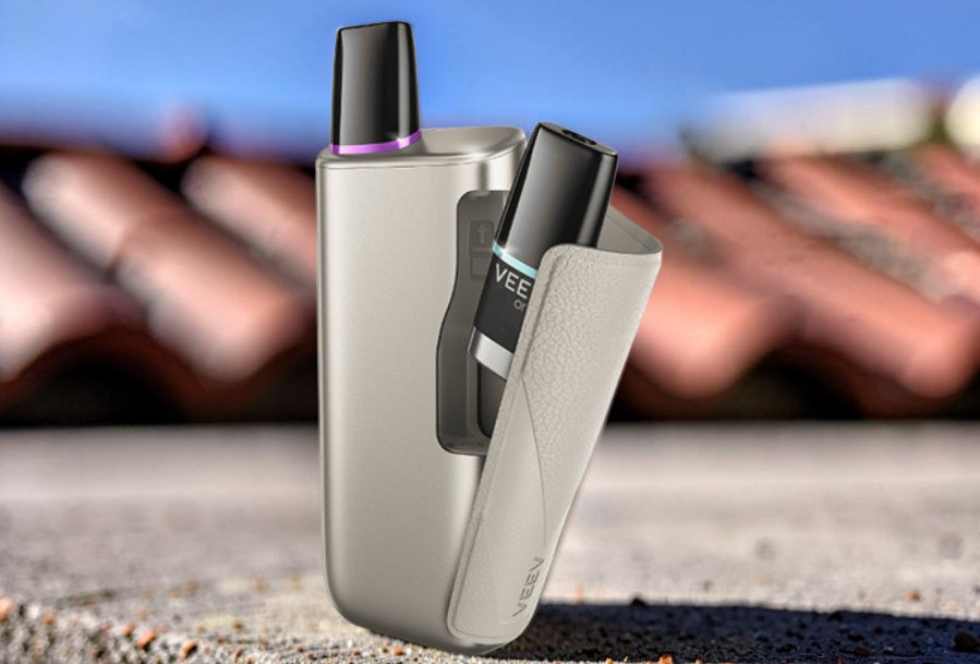 A gold-finished Veev One Plus pod system kit with its built-in storage compartment slid open. A pre-filled VEEV ONE pod is loaded in the active heating chamber while an extra backup pod sticks out of the storage slot.